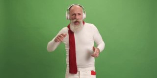 一位看起来像圣诞老人的老人用智能手机听音乐、跳舞、唱歌。在绿色的背景上。一个人。