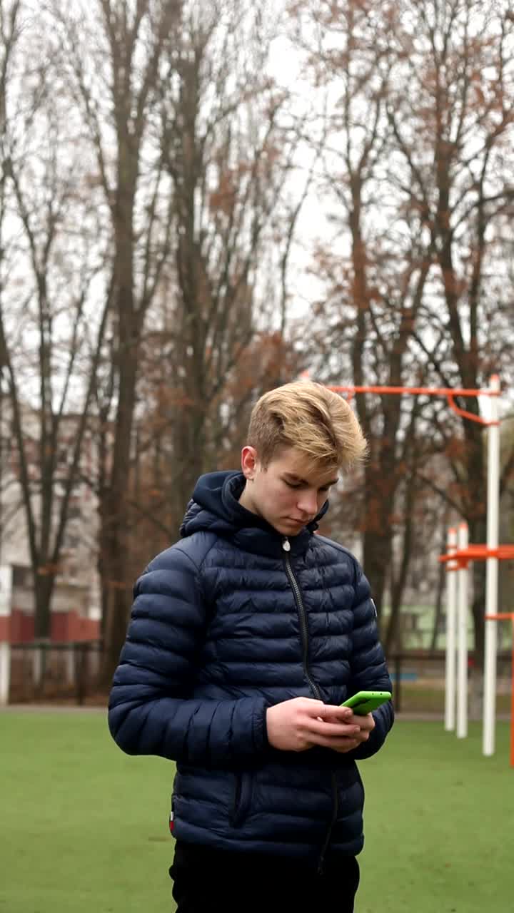 垂直视频，男子阅读在线新闻，并在一个社交网络上用智能手机写短信在运动场。在秋天的操场上，穿着蓝色夹克的年轻帅哥正在用手机。