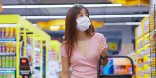 亚洲妇女戴着口罩，拿着购物篮在超市购物。在冠状病毒危机或新冠肺炎爆发期间，女孩在货架上挑选、寻找食物。