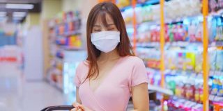 亚洲妇女戴着口罩，拿着购物篮在超市购物。在冠状病毒危机或新冠肺炎爆发期间，女孩在货架上挑选、寻找食物。