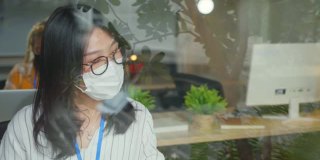 年轻创意设计师代理在防护口罩中为保护大流行病毒一起工作笔记本电脑。亚洲商人和女商人讨论和咨询新的正常生活方式