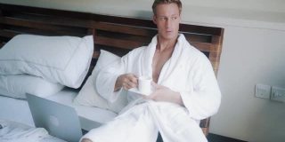 穿着浴袍的年轻人在床上喝咖啡，旁边是笔记本电脑