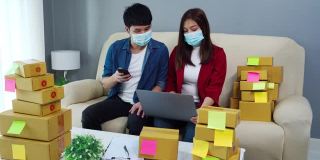 夫妇企业家用笔记本电脑和智能手机准备快递给客户在家里的办公室，人们戴着口罩保护冠状病毒(covid-19)大流行