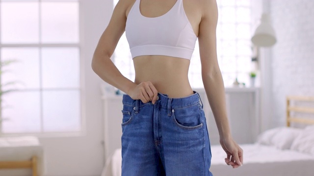 年轻苗条的女人穿着大牛仔裤在家里展示她的减肥结果