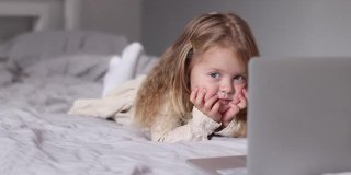 漂亮的金发小女孩穿着裙子在灰色的床上用笔记本电脑看卡通片。早上的例行公事，童年的，蹒跚学步的，女儿的。FullHD镜头