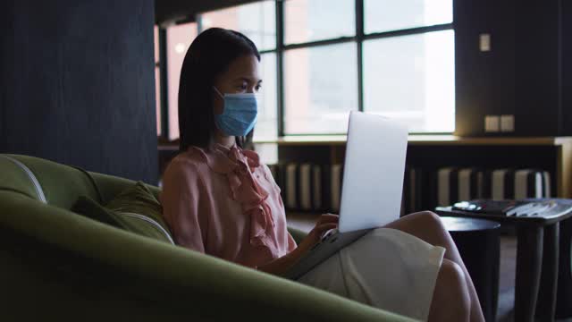亚洲妇女戴着口罩，坐在沙发上使用笔记本电脑在现代办公室