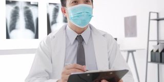 严重的亚洲男医生戴着防护口罩，用剪贴板与女病人在医院的办公室里传递好消息，谈论结果或症状。