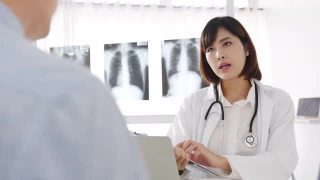 年轻的亚洲女医生正在用剪贴板传递好消息，谈论结果或症状与男性病人坐在健康诊所的桌子上。视频素材模板下载