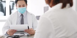 严重的亚洲男医生戴着防护口罩，用剪贴板与女病人在医院的办公室里传递好消息，谈论结果或症状。