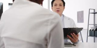 严肃的亚洲男医生正在用剪贴板与女病人坐在医院办公室的桌子前，传递好消息，谈论结果或症状。