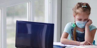 女孩在网上上课，做作业，看着窗外。网络学校远程学习隔离一个无聊的小女孩一边在笔记本上写字，一边看着笔记本电脑。