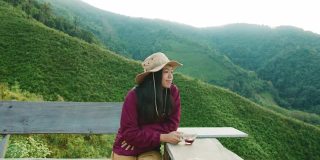 亚洲女人拿着咖啡杯和喝咖啡在山顶的背景。慢旅行的生活方式与浸咖啡的概念。4 k慢动作。
