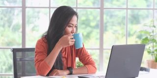 年轻美丽的亚洲女人喝咖啡，与笔记本电脑在家里办公，人与技术，在家里工作，在线教育