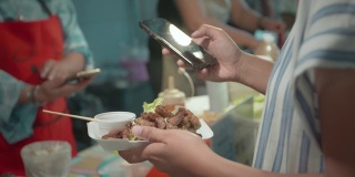 一名亚洲女子在夜市街头买了烤猪肉后，用智能手机扫描条形码付款。泰国的街头小吃。