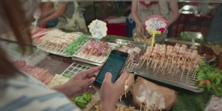 一名亚洲女子在夜市买了猪肉串、肉串和蔬菜串后，用智能手机扫描条形码付款。泰国的街头小吃。