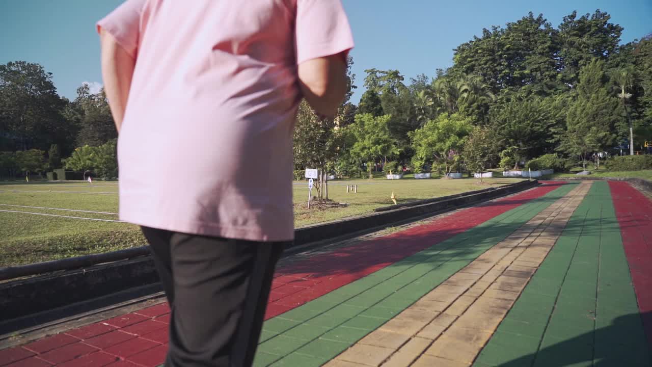 在一个阳光明媚的日子里，亚洲老年肥胖女性在公园的跑道上慢动作跑步。退休生活方式的活动。医疗动机，脉搏血压跟踪，后视镜