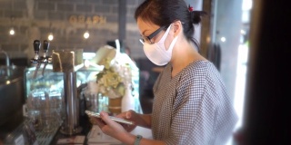 两名亚洲妇女戴着防护口罩在咖啡店用智能手机付款