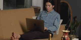 亚洲女性在网上工作，与朋友或客户交流，坐在沙发上，在家里的暗室里用笔记本电脑