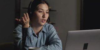 一个年轻的亚洲女人的特写微笑在耳机与麦克风工作，与朋友聊天或客户在线视频会议，在一个黑暗的房间在家里使用笔记本电脑