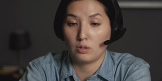 一个年轻的亚洲女人的特写在耳机与麦克风与客户在线视频会议，使用笔记本电脑在一个黑暗的房间在家里