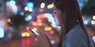 微笑的年轻亚洲女性使用智能手机发短信或网上购物的夜晚，在城市的路灯照明。漂亮女孩享受和享受城市夜生活的乐趣。