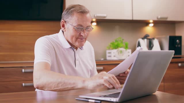 快乐的老人坐在厨房的椅子上，用笔记本电脑购买网上网站，凭证和银行信用卡预订酒店计划度假，老人在网上学习支付系统