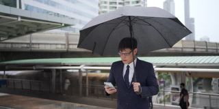 自信的亚洲商人办公室职员在雨中拿着黑色雨伞使用智能手机。帅哥商务人士工作在城市办公区在恶劣的天气。
