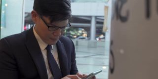 亚洲商人办公室职员坐在咖啡店的窗口使用智能手机上网商务在线工作或短信，同时等待在咖啡馆的伙伴关系。商业和技术概念。