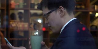 亚洲商人办公室职员坐在咖啡店的窗口边喝咖啡，使用智能手机和互联网在网上工作的公司业务或短信在晚上。商业和技术概念。