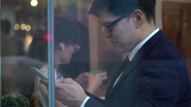 亚洲商人办公室职员坐在咖啡店的窗口使用智能手机上网商务在线工作或短信，同时等待在咖啡馆的伙伴关系。商业和技术概念。