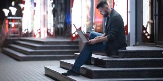 晚上，一个留着胡子的英俊男子坐在大街上打字，腿上放着笔记本电脑。想着远处的东西。自由工作的概念。聊天、发电子邮件、搜索或约会。