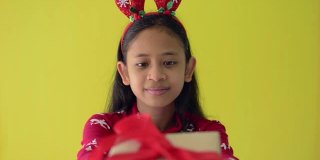 快乐的长头发漂亮的女孩穿着红毛衣和戴着驯鹿鹿角头带收到了一个圣诞礼物盒孤立在黄色的背景。
