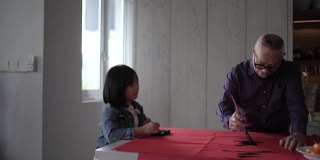 亚洲华人祖父和孙女在中国新年庆祝期间练习书法，家里装饰一张红色的纸