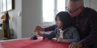 亚洲华人祖父和孙女在中国新年庆祝期间练习书法，家里装饰一张红色的纸