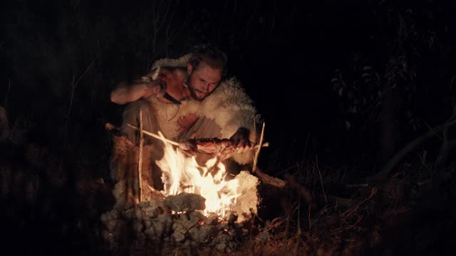 一个披着兽皮的大胡子男人，晚上在乡下，在火边休息，吃苹果。晚上在火边吃东西的野蛮人