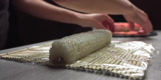 在正宗的日本餐厅工作时，匿名厨师的手与竹垫子滚动新鲜寿司，厨师的手在竹垫子上滚动寿司卷的特写。寿司制作过程。米饭，紫菜，鳄梨，螃蟹，红鱼子酱。卷的