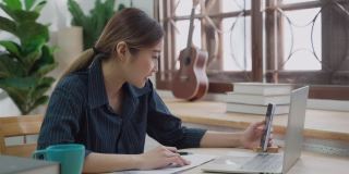 快乐的年轻亚洲妇女微笑与手机，同时使用笔记本电脑在家里工作