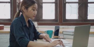 快乐的年轻亚洲妇女微笑与手机，同时使用笔记本电脑在家里工作