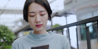 十几岁的亚洲短发女人坐在智能手机微笑，高兴地选择网上购物。假期感觉轻松，快乐。在公共场所使用5G技术和免费Wi-Fi