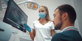 牙科医生戴着手套在牙科诊所的电子屏幕上展示牙齿全景x光片，背景为光，配有医疗设备。微笑健康牙齿概念。