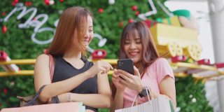 两位快乐的亚洲年轻女性在百货商店的户外购物在销售季节在年底。女孩们拿着购物袋，快乐地走着，微笑着一起买东西