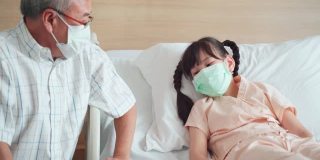 亚洲小女孩戴着口罩躺在康复室的床上，父亲来到康复室探望并照顾她的女儿。