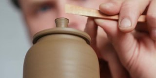 一个男性陶工的特写镜头，从一个陶工的车轮上的一个陶土茶壶的壶盖的把手中去除多余的粘土。陶工制作粘土制品