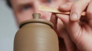 一个男性陶工的特写镜头，从一个陶工的车轮上的一个陶土茶壶的壶盖的把手中去除多余的粘土。陶工制作粘土制品视频素材模板下载