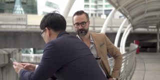 4K两个商务人士白种人与亚洲商人同事伙伴关系讨论商业项目计划与使用数字平板电脑在城市。商务会议、技术、并购概念。