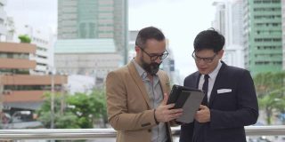 4K两个商务人士白种人与亚洲商人同事伙伴关系讨论商业项目计划与使用数字平板电脑在城市。商务会议、技术、并购概念。