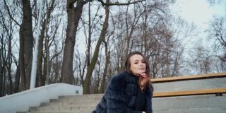 美丽的模特坐在公园里的肖像。美丽的年轻女子在黑裘皮大衣摆姿势在相机户外。