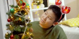 快乐的亚洲男孩穿着绿色的毛衣和红色的发带装饰圣诞树在晚上的时间在家里，节日的概念。