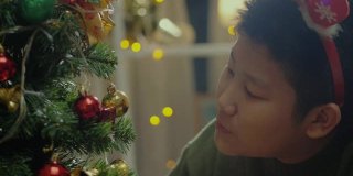 快乐的亚洲男孩穿着绿色的毛衣和红色的发带装饰圣诞树在晚上的时间在家里，节日的概念。