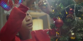 快乐的亚洲男孩穿着红色的毛衣和发带装饰圣诞树在晚上的时间在家里，节日的概念。